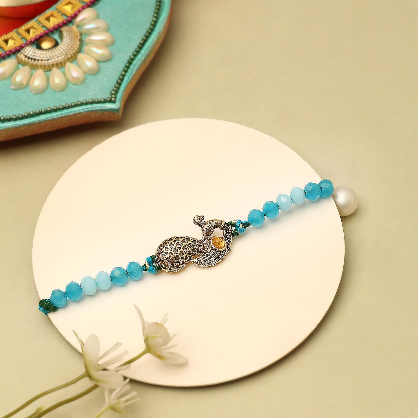 A vibrant peacock charm adorns a rakhi, accompanied by a bowl of red powder, all set on a beautifully arranged table.