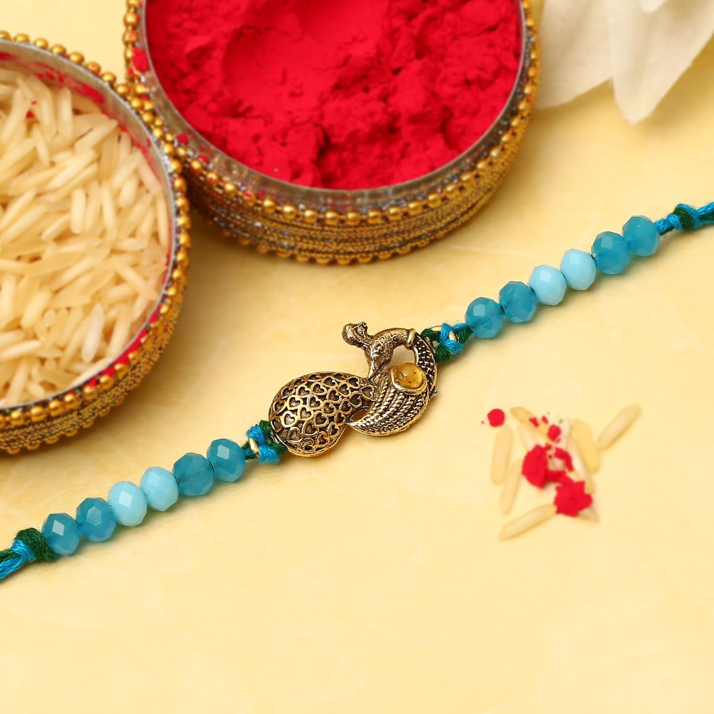 A peacock charm sits on a rakhi next to a bowl of red powder on a table, creating a festive and colorful display.