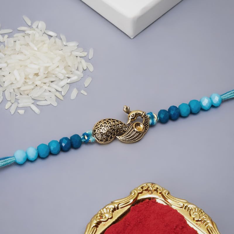 A peacock charm is placed on a rakhi, with a bowl of red powder beside it, creating a colorful scene on the table.