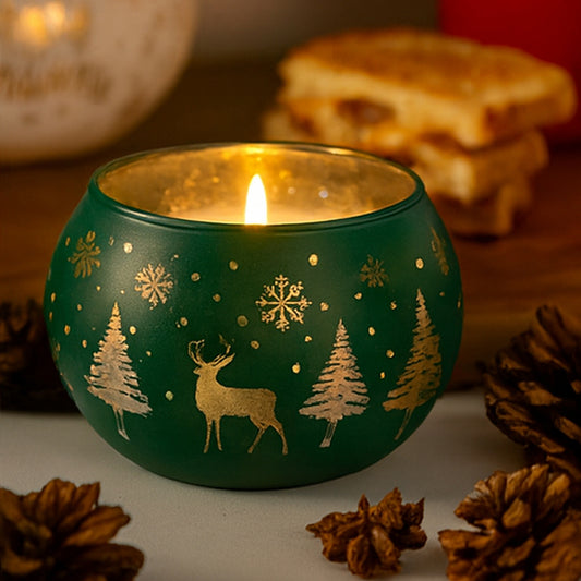 Green candle holder with gold reindeer and tree designs, lit candle inside, surrounded by pine cones and a blurred background.