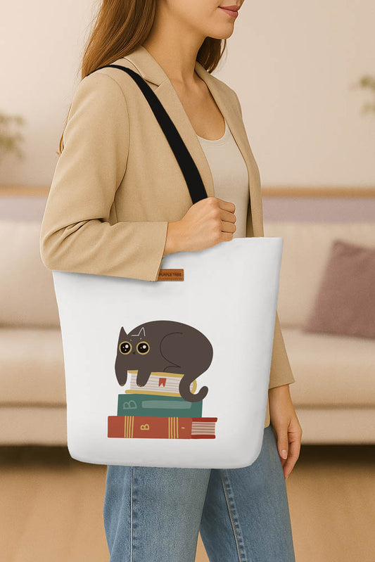 A tote bag adorned with a cat sitting atop a pile of books, emphasizing a love for reading and feline companionship.