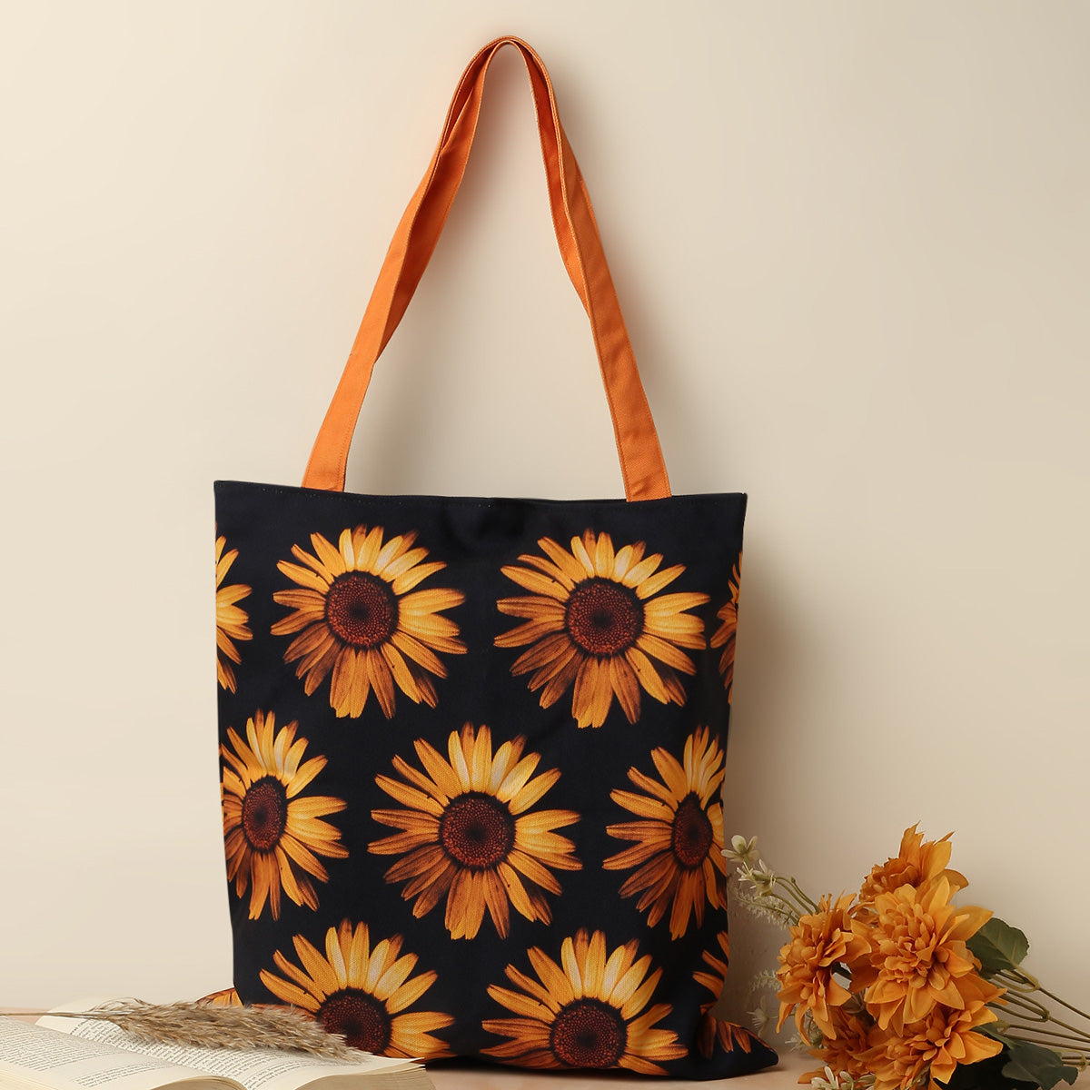 A stylish tote bag in black color and orange sunflower design on it with orange color handles adorned with beautiful bunch of flowers beside on a table and beige background