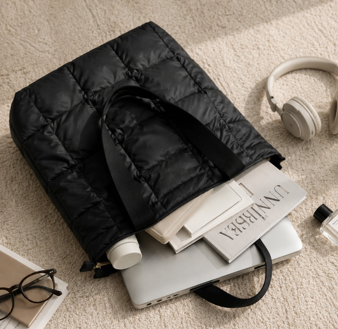 Black puffer bag on a stack of books with headphones and glasses nearby on a beige carpet.