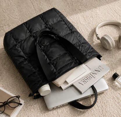 Black puffer bag on a stack of books with headphones and glasses nearby on a beige carpet.
