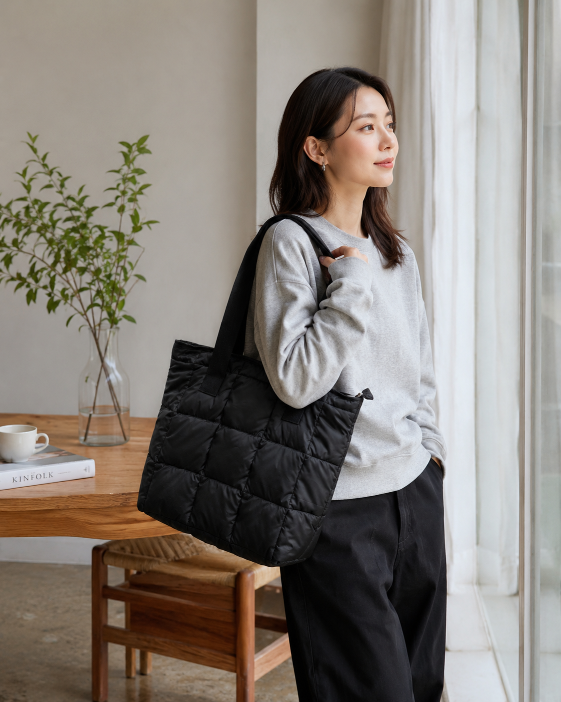 Woman holding a black quilted tote bag in a room with a wooden table and plant.