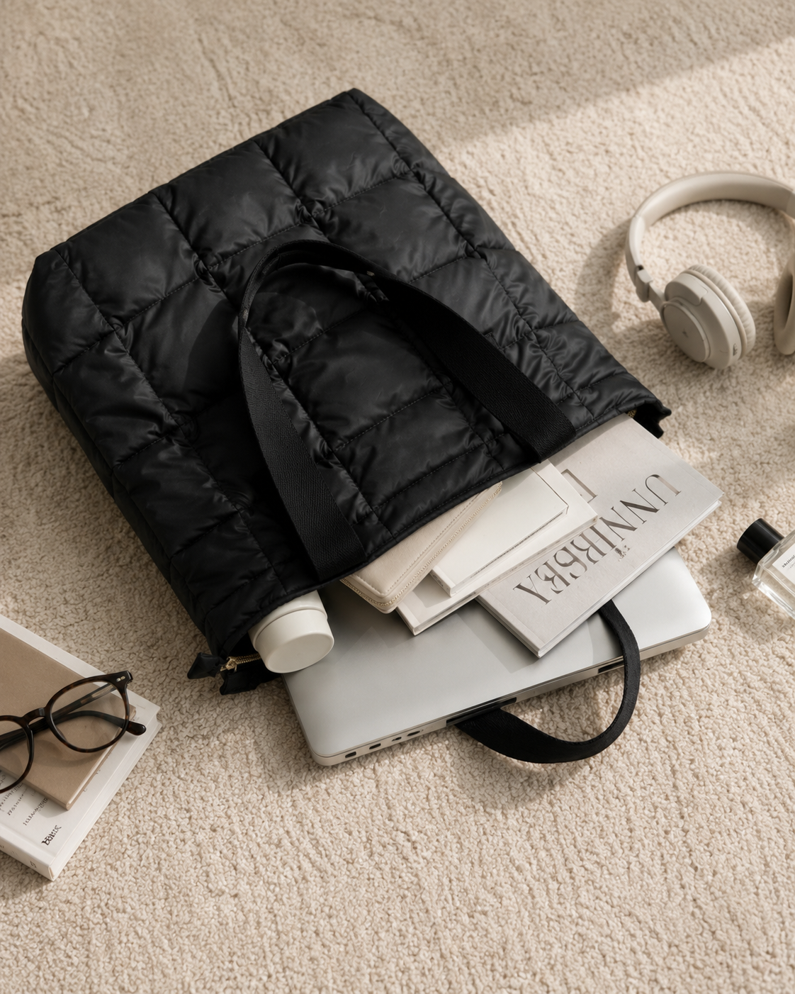 Black quilted tote bag on a stack of books with a laptop, glasses, and headphones on a beige carpet.