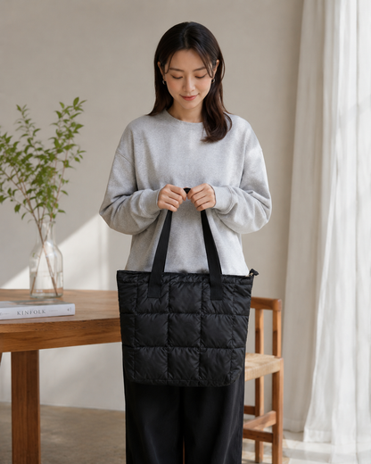 Woman holding a black puffer quilted tote bag in a room with a wooden table and plant.
