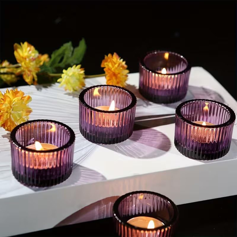 Set of purple candle holders with lit candles on a white surface with yellow flowers in the background.