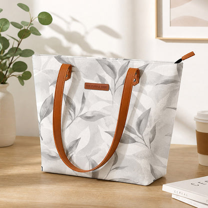 White tote bag with leaf pattern and brown handles on a wooden surface.