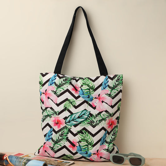A tote bag with a tropical floral print on a black and white chevron pattern, with sunglasses and magazines beside it.