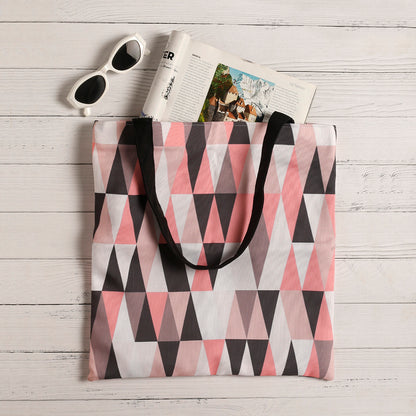 A stylish tote bag with a pink, grey and black triangle pattern, resting on a white wooden surface with sunglasses and a magazine partially tucked inside.