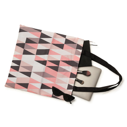 A stylish pink, black, and white geometric patterned tote bag with a black strap, partially opened to reveal a laptop, makeup brushes, and a pair of black sunglasses.