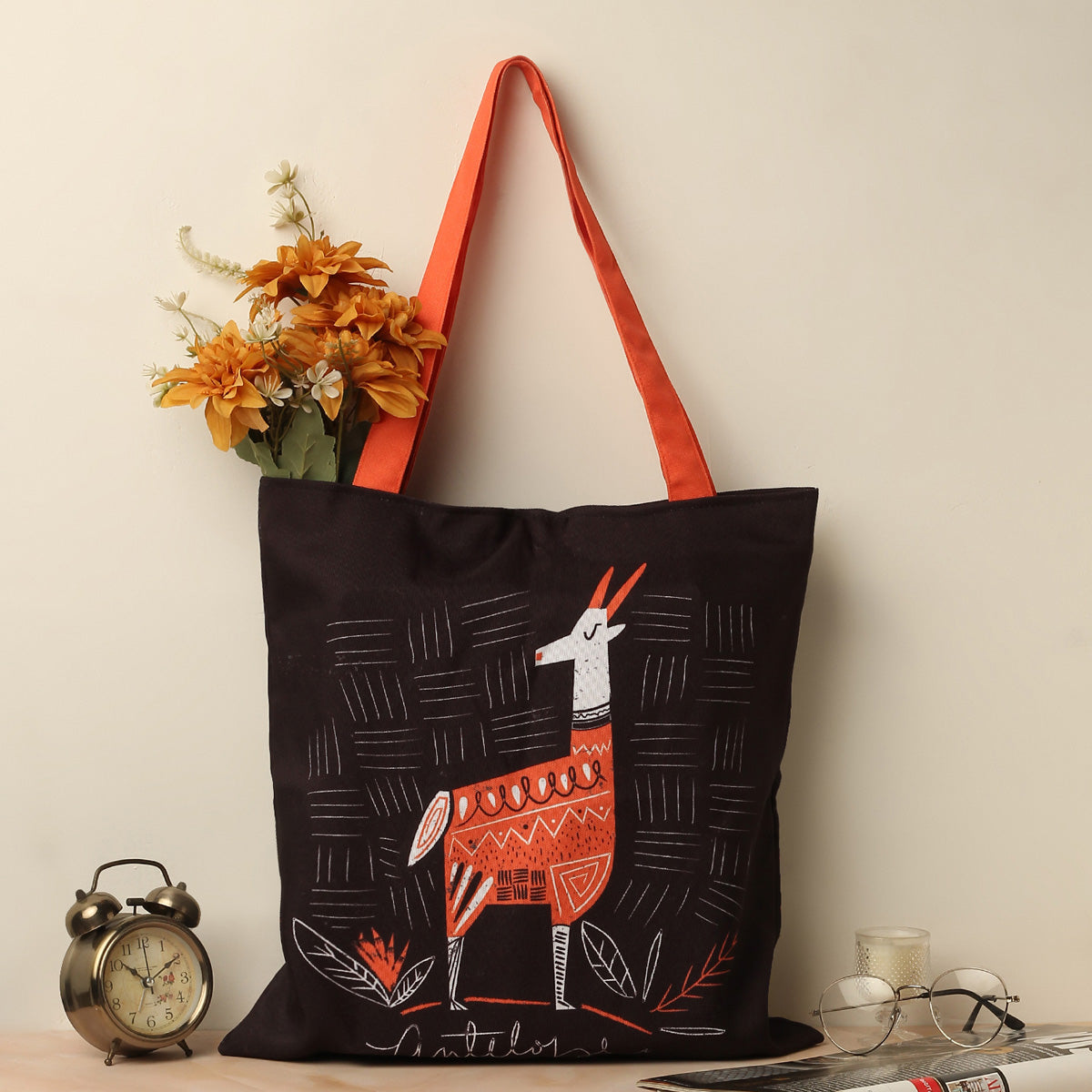 A black tote bag with a orangish strap, featuring a white and orange Deer design, placed on a table next to an alarm clock, glasses, and a candle holder. There are orange flowers in the bag.