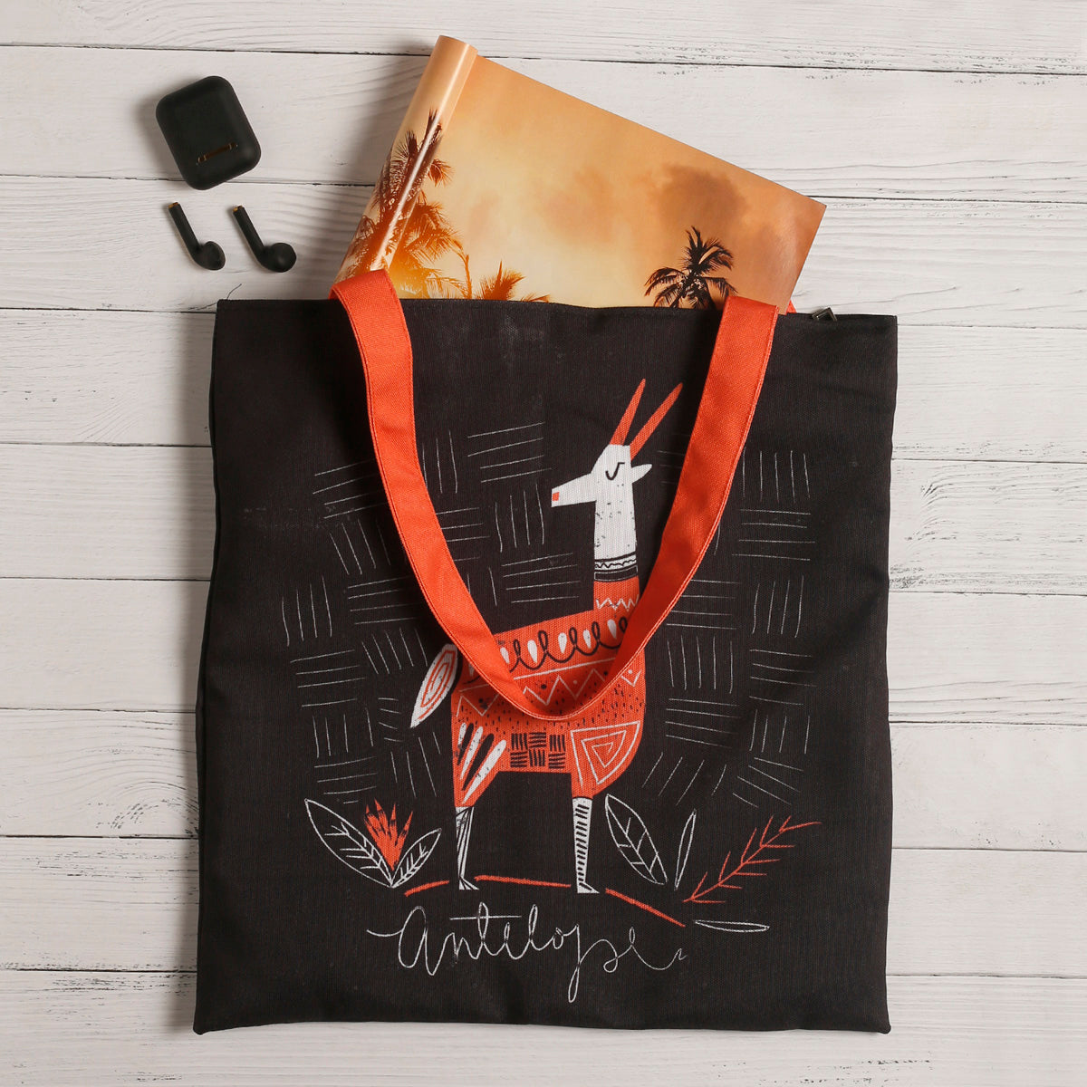 Black tote bag with orange handles and tribal print design featuring a deer, plants.  A pair of wireless earbuds and a charging case are placed beside it, along with a notebook with a sunset and palm tree cover peeking out from the bag. The items are on a white wooden surface.