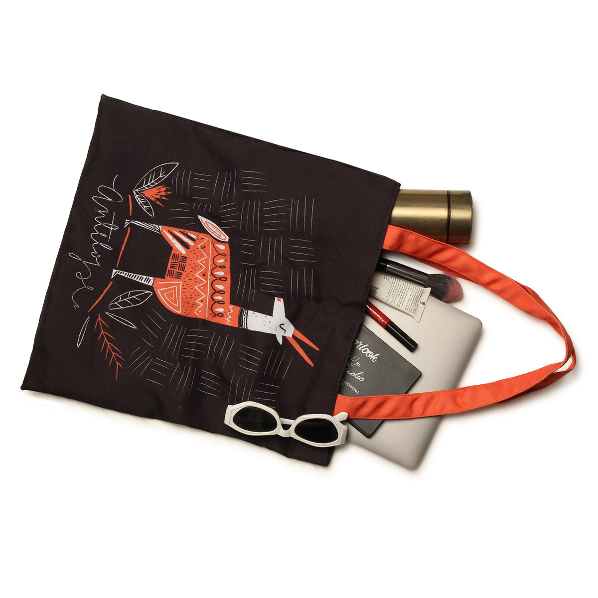A black tote bag with a red and white tribal deer design, an orange strap, and various items inside including a gold water bottle, sunglasses, a notebook, and makeup brushes.