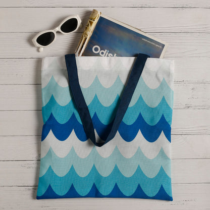A patterned tote bag with blue wave design, a pair of white sunglasses, and a travel book titled "Odisea" on a white wooden surface.