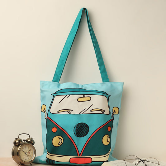 A teal tote bag with a printed design of a vintage van on it, placed next to a pair of glasses and an old-fashioned alarm clock on a table.