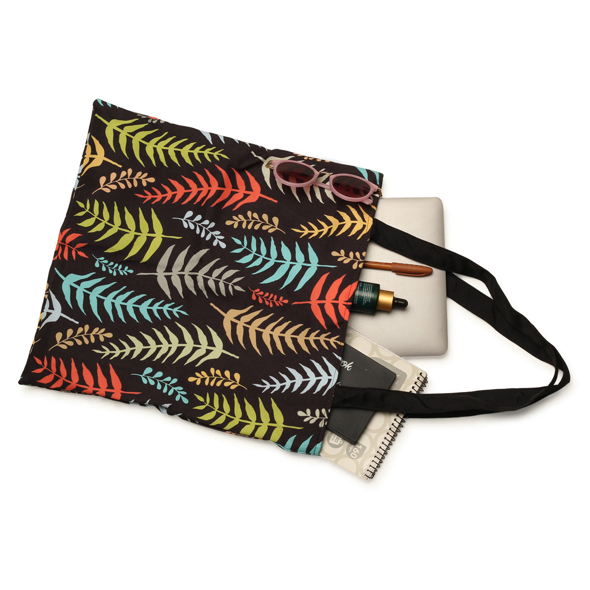 A black tote bag with colorful leaf patterns, a pair of sunglasses, a laptop, a notebook, and two bottles of essential oils placed on top.