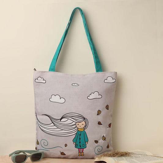 A light grey tote bag with teal straps featuring an illustration of a girl with long hair blowing in the wind surrounded by clouds and fallen leaves, next to a pair of sunglasses and an open book on a beige background.