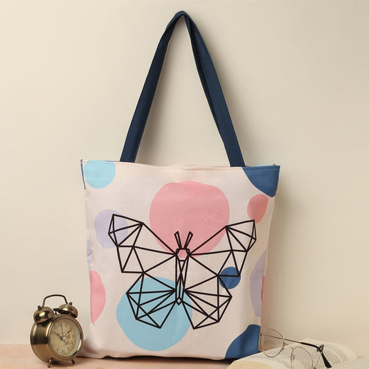 A canvas tote bag with a geometric butterfly design and pastel-colored polka dots, placed next to a vintage alarm clock and a pair of eyeglasses.