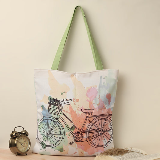 A white tote bag with green handles featuring a colorful watercolor background and a black line drawing of a vintage bicycle with a basket of flowers. The bag is standing against a pale yellow background, next to a vintage gold alarm clock and an open book with a feather.