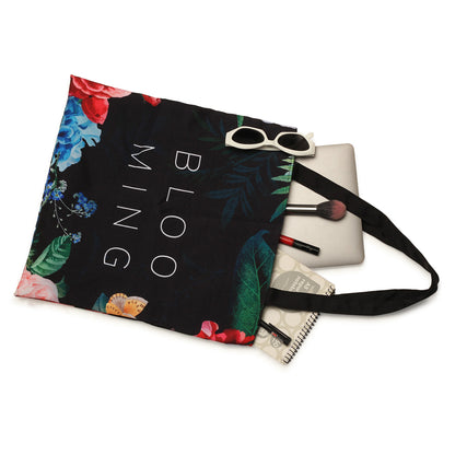 A floral tote bag labeled "blooming" containing sunglasses, a laptop, a notebook, and makeup brushes, displayed on a white background.