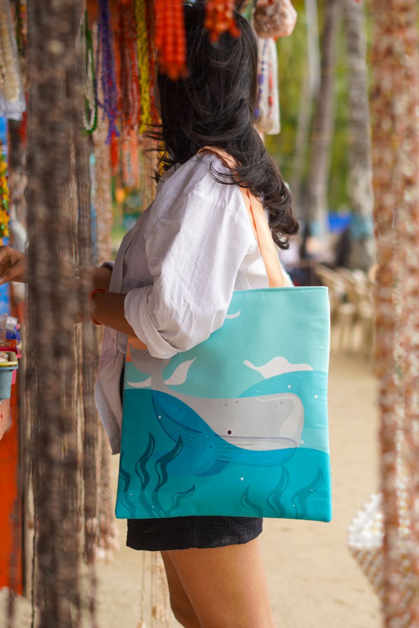 A woman with long dark hair wearing a white shirt and a black skirt, holding a large turquoise tote bag with a printed design of a whale on it, standing in front of a market stall with colorful beaded necklaces.