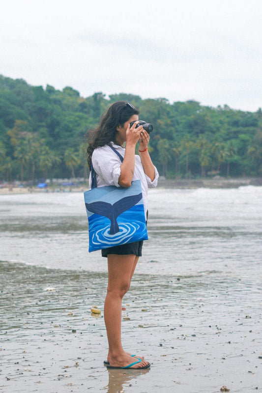 A person standing on a sandy beach holding binoculars to their eyes, wearing a white shirt, black shorts, blue flip-flops, with a shoulder bag featuring a whale tail design. There are palm trees and a hazy sky in the background.