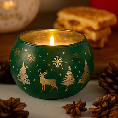 Green candle holder with gold reindeer and tree designs, lit candle inside, surrounded by pine cones and a blurred background.