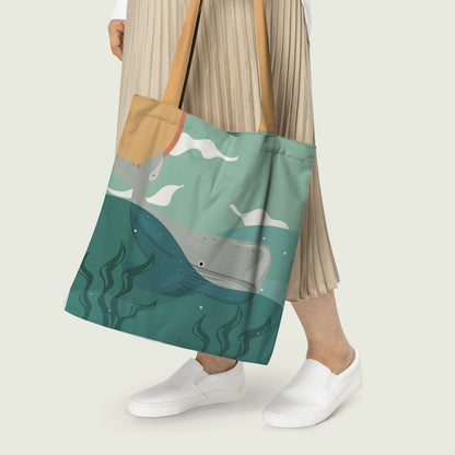Person holding a tote bag with a design of a whale and waves on a teal background.