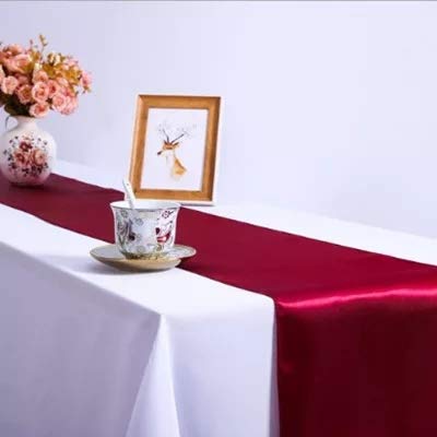 A maroon satin table runner is laid out on a white table with a cup and a vase containing flowers on top of it, with a picture frame and a decorative flower vase in the background.